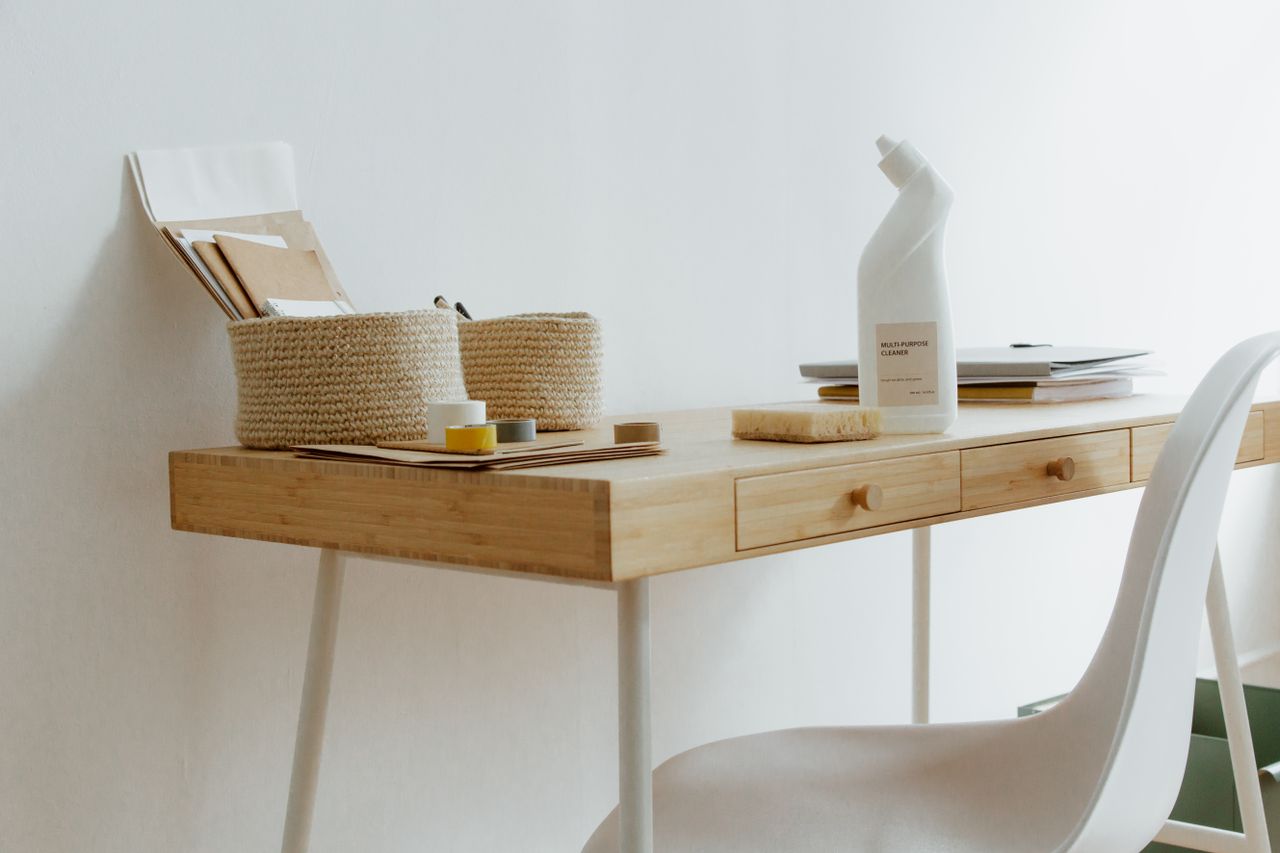 A tidy, well-arranged home office desk featuring cleaning products, papers, and storage baskets.